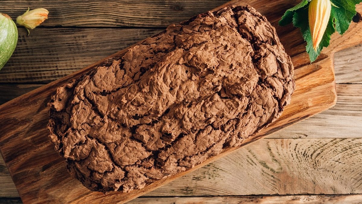 Draufsicht: Ein Kuchen auf einem Holzbrett.. Die Oberfläche ist nicht glatt, sondern rau. Es handelt sich um einen Schokoladen-Zucchini-Kuchen.