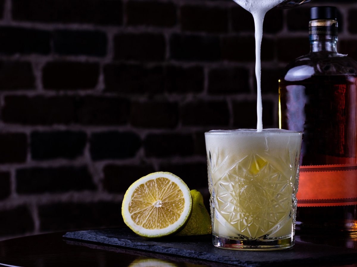 Whiskey Sour oder Boston Sour beim Eingießen in Old Fashioned Glas vor auf Holztisch Backsteinwand. Daneben Zitronen und Whiskeyflasche. Forntalansicht.