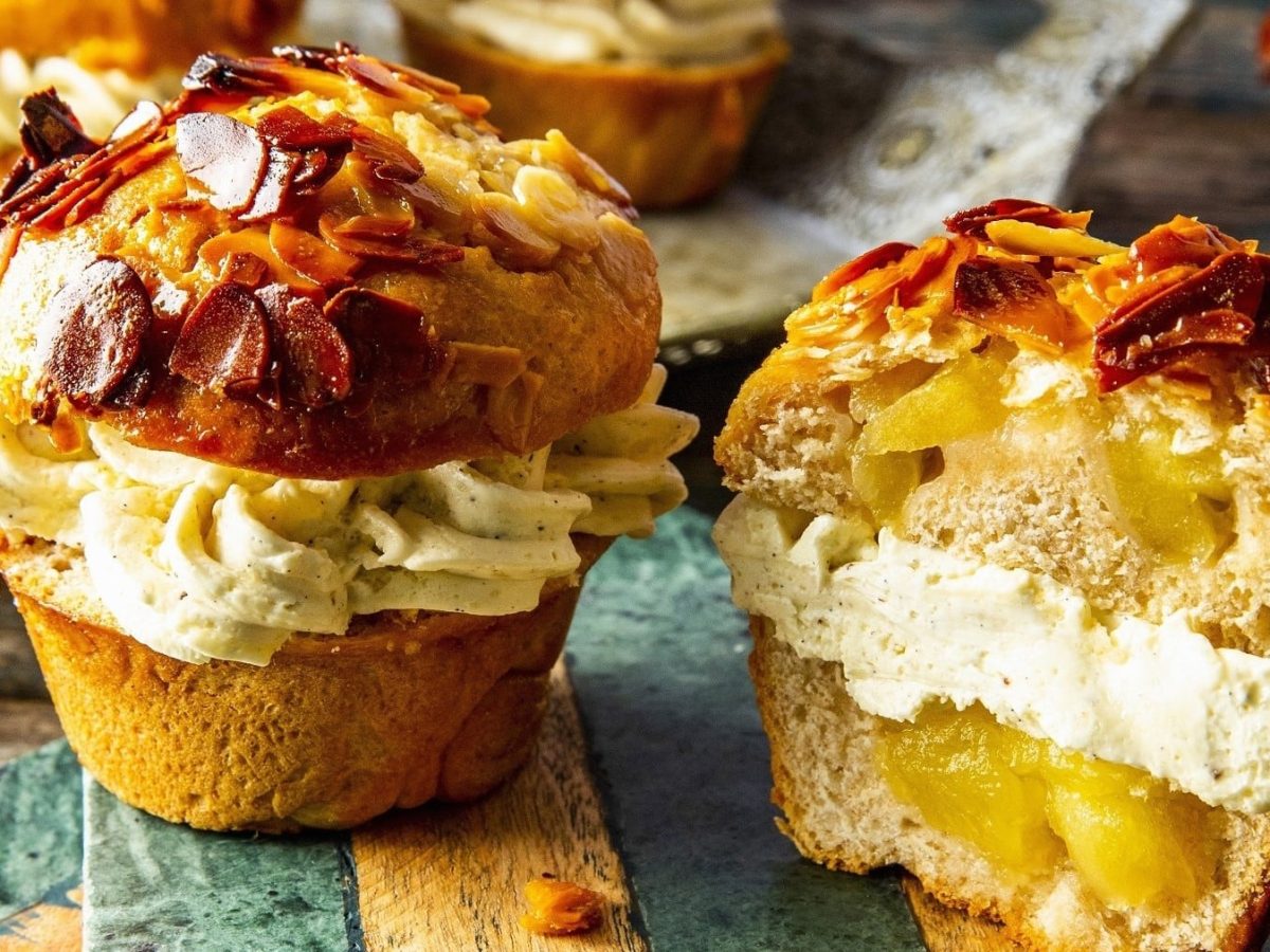 Apfel-Bienenstich-Muffins, angeschnitten serviert auf einem einfachen, bunten Holzbrett, garniert mit karamellisierten Mandeln.