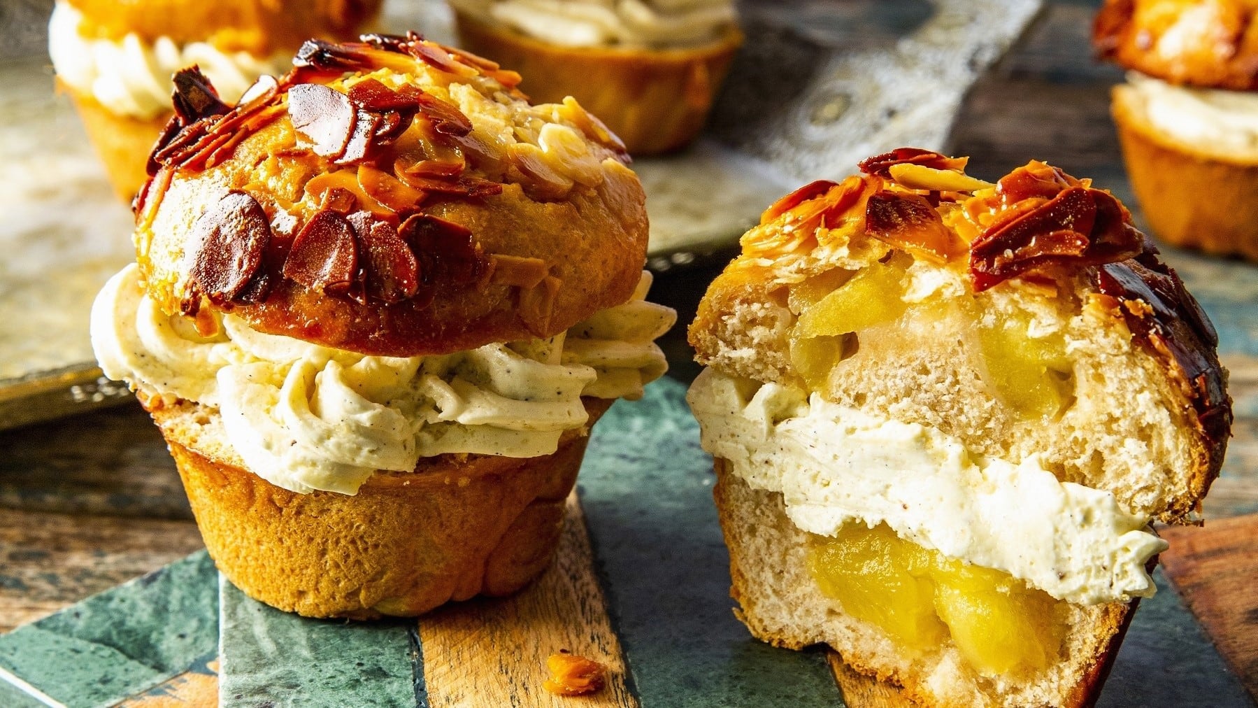 Apfel-Bienenstich-Muffins, angeschnitten serviert auf einem einfachen, bunten Holzbrett, garniert mit karamellisierten Mandeln.