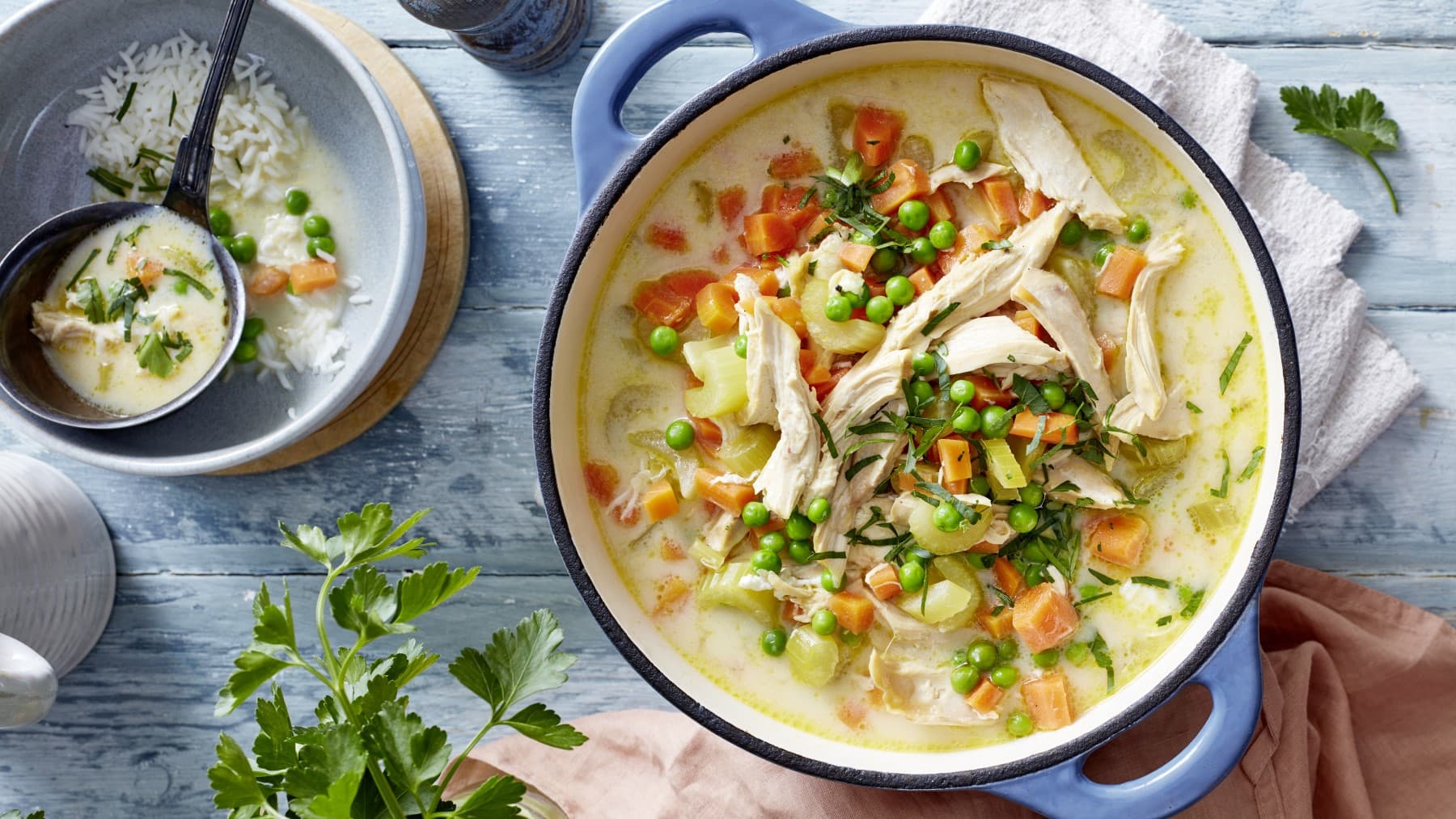 Griechische Hühnersuppe mit Petersilie im Topf neben einer Schüssel mit Reis und einer Kelle Suppe vor blauem Hintergrund,
