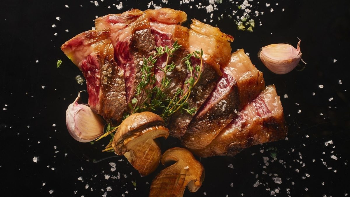 Rindfleisch-Tagliata mit Pilzen und gebackenem Knoblauch, auf einer schwarzen Schieferplatte serviert und mit frischen Kräutern und grobem Salz garniert.