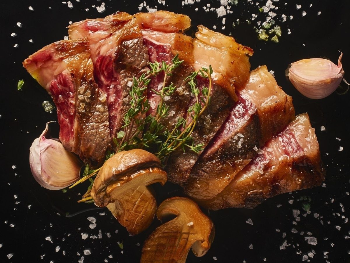Rindfleisch-Tagliata mit Pilzen und gebackenem Knoblauch, auf einer schwarzen Schieferplatte serviert und mit frischen Kräutern und grobem Salz garniert.