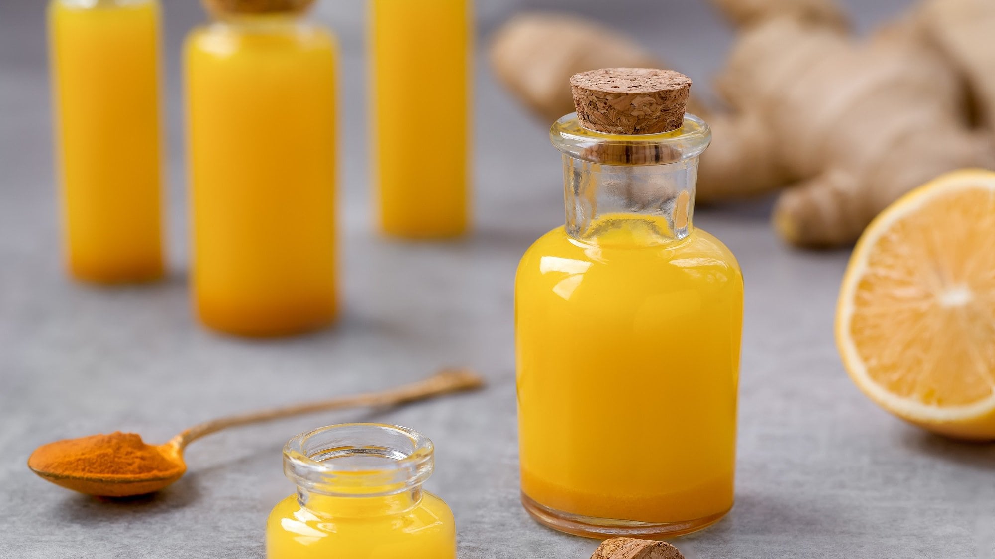 Mehrere Flaschen Ananas-Ingwer-Shots, drumherum ein Löffel Kurkuma, Ingwerwurzeln und eine Orange.