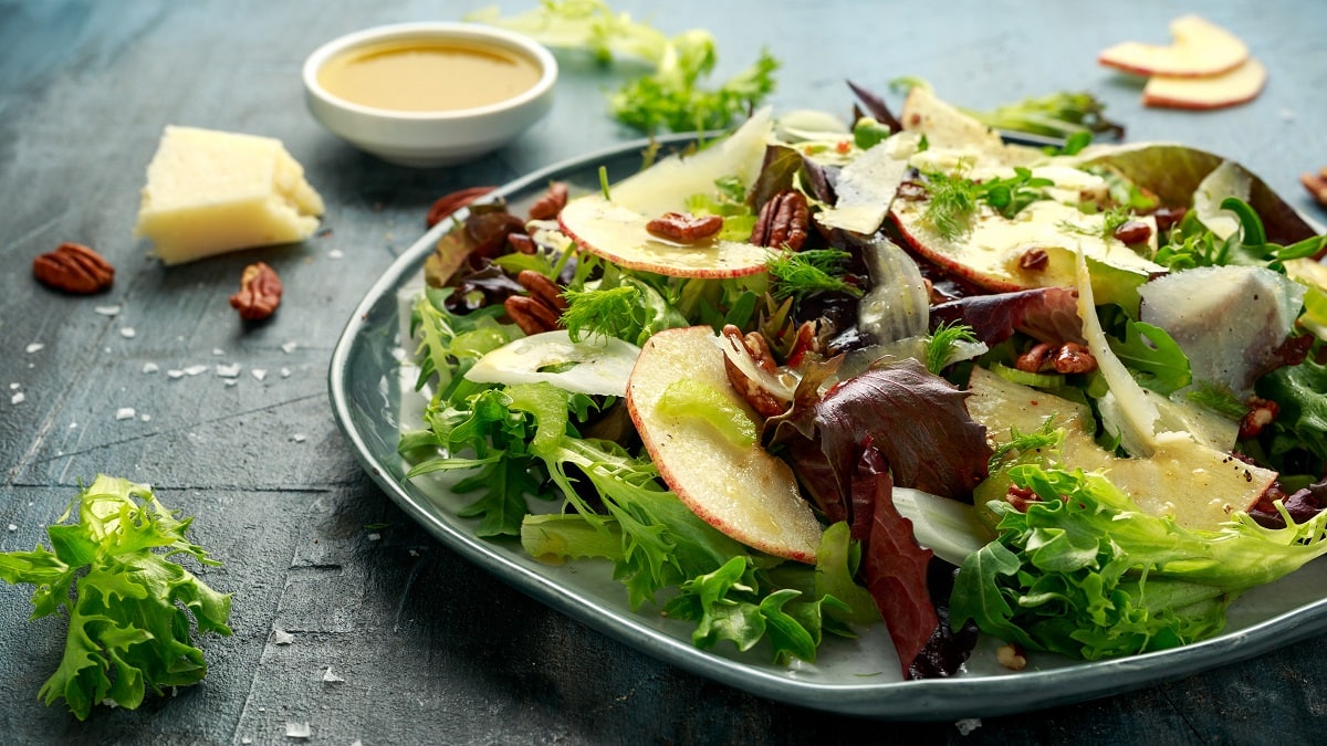 Frontalansicht: EIn Teller Apfel-Fenchel-Salat steht auf einem festen Untergrund. Drumherum liegen ein Stück Käse, eine Schale mit einer Flüssigkeit und Salatblätter.