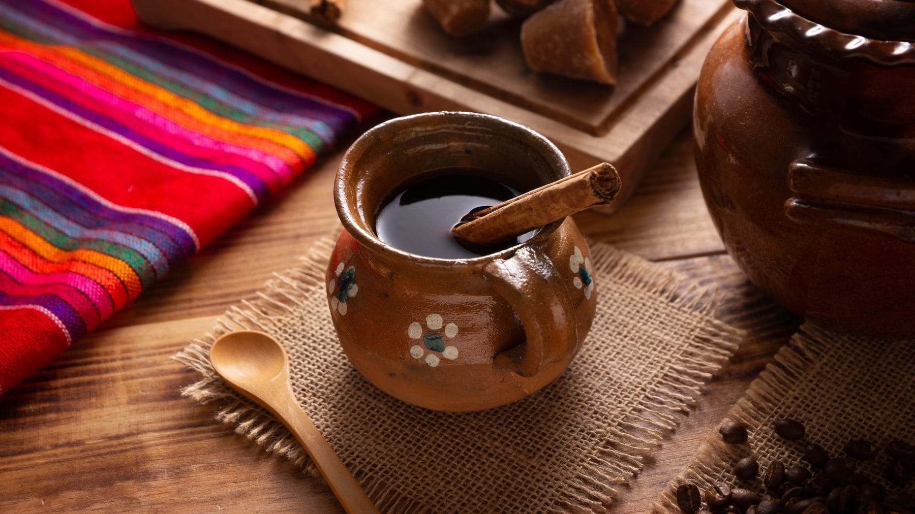 Café de Olla traditioneller mexikanischer Kaffe in Tongefäß mit Zimtstange auf Stofftuch. Daneben traditionelles mexikanisches Tischtuch, brauner Zucker, eine Tonkanne und Zimtstangen. Draufsicht.