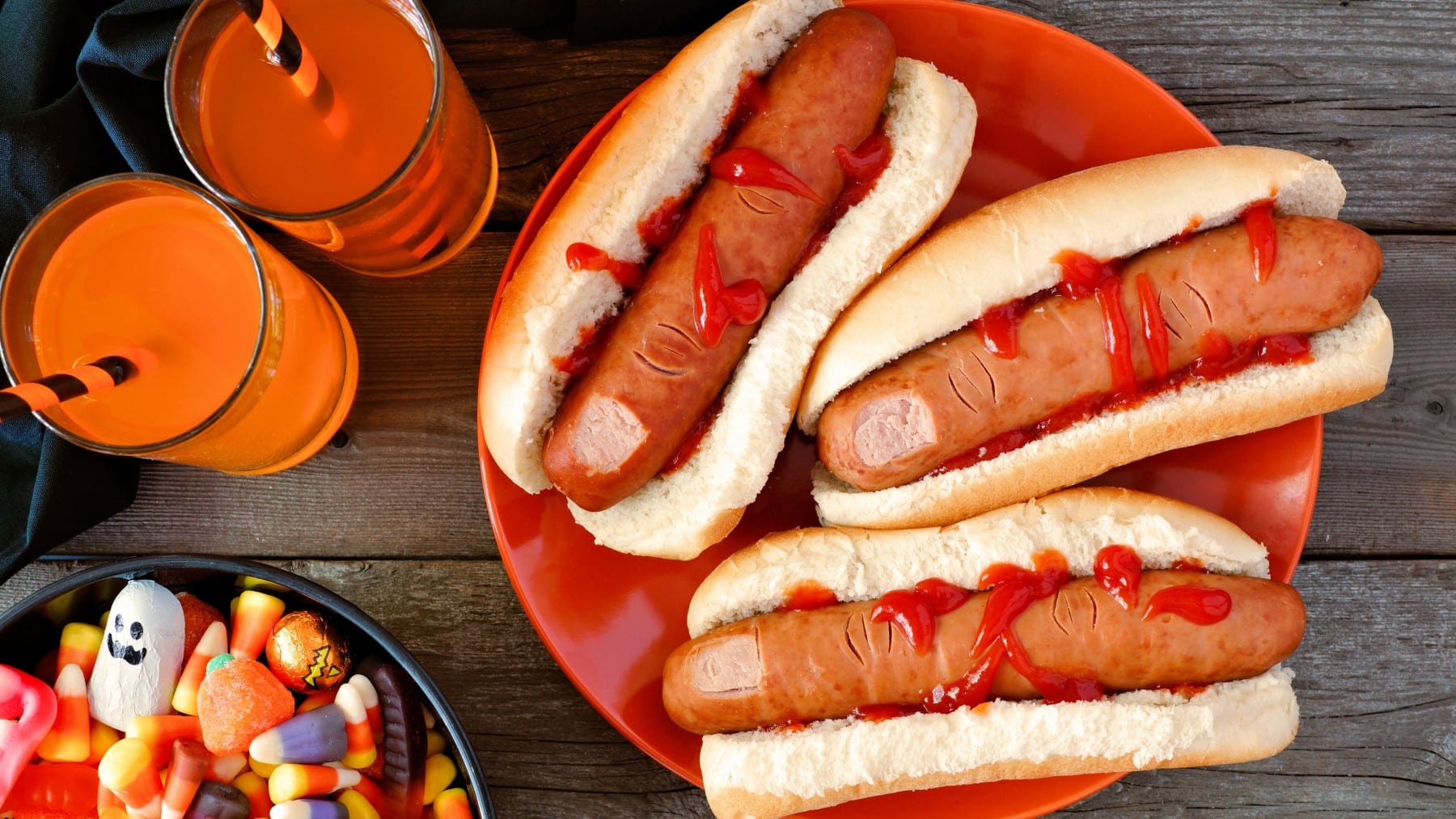 Drei Finger Hot Dogs mit Ketchup auf einem orangenen Teller, neben orangenen Getränken und Süßigkeiten vor grauem Holzhintergrund.