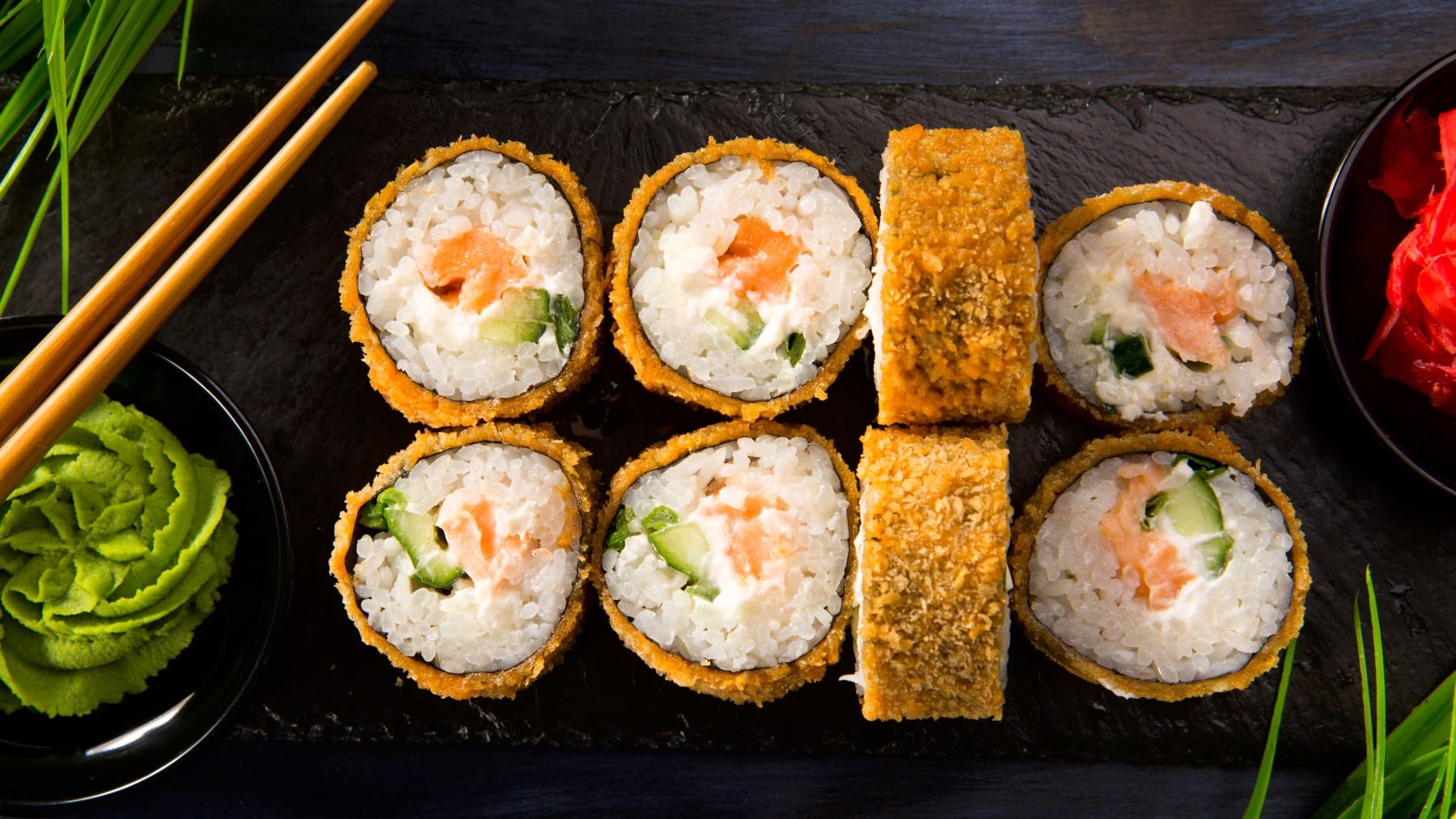 Acht Fried Sushi Röllchen vor dunklem Hintergrund neben Wasabi und Stäbchen.