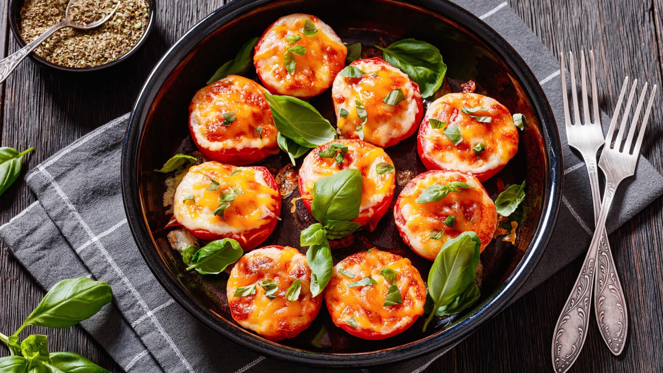 Gebackene Tomaten mit dreierlei Käse auf schwarzem Teller mit Basilikum auf grauem Tuch und Tisch. Oben Schale mit Oregano. Daneben Besteck und Basilikum. Draufsicht.