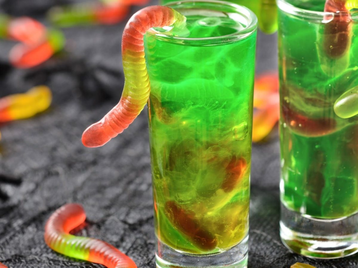 Shotgläser mit grünen Halloween Jello Shots mit Gummiwürmern auf schwarzer Fläche mit mehr Würmern im Hintergrund.