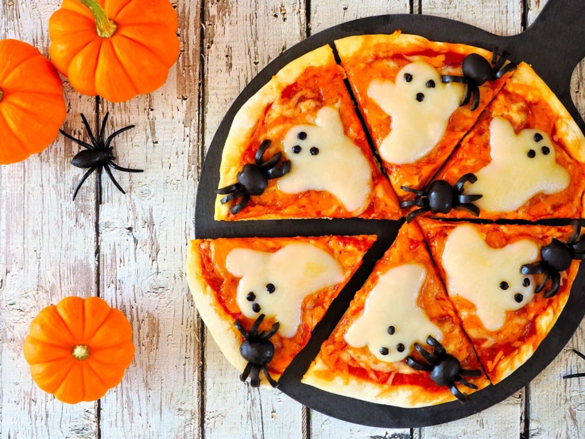 Halloween-Pizza mit Käse in geisterform und Oliven-Spinnen vor Holzhintergrund neben Halloween Deko.