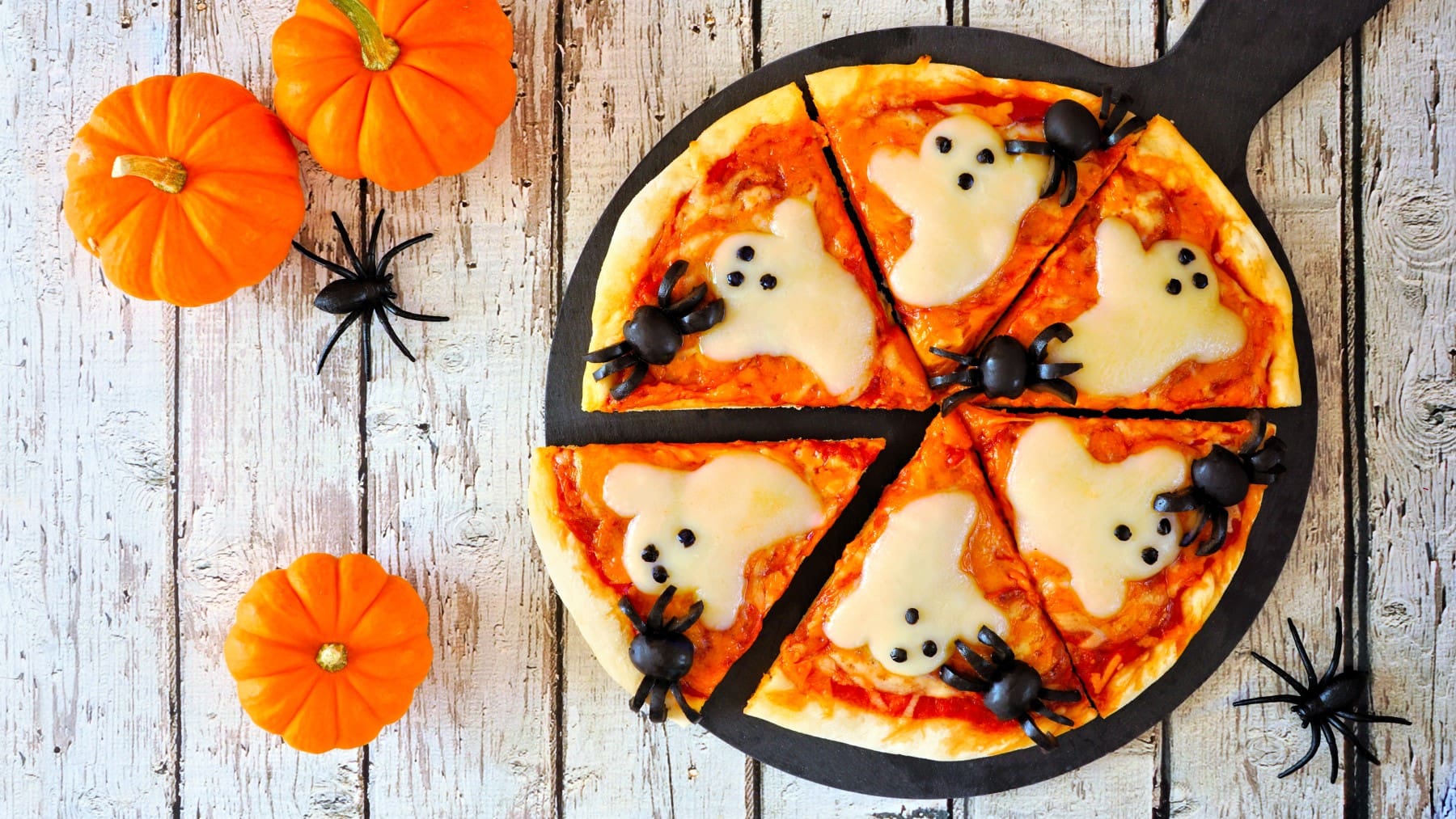 Halloween-Pizza mit Käse in geisterform und Oliven-Spinnen vor Holzhintergrund neben Halloween Deko.