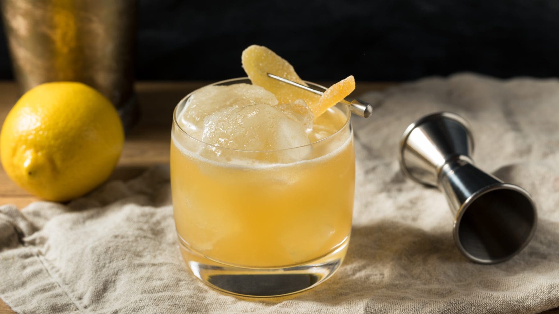 Penicillin Cocktail mit kandiertem Ingwer auf Tuch mit Barmaß und Zitrone. Im Hintergrund Cocktailshaker. Frontalaufnahme.