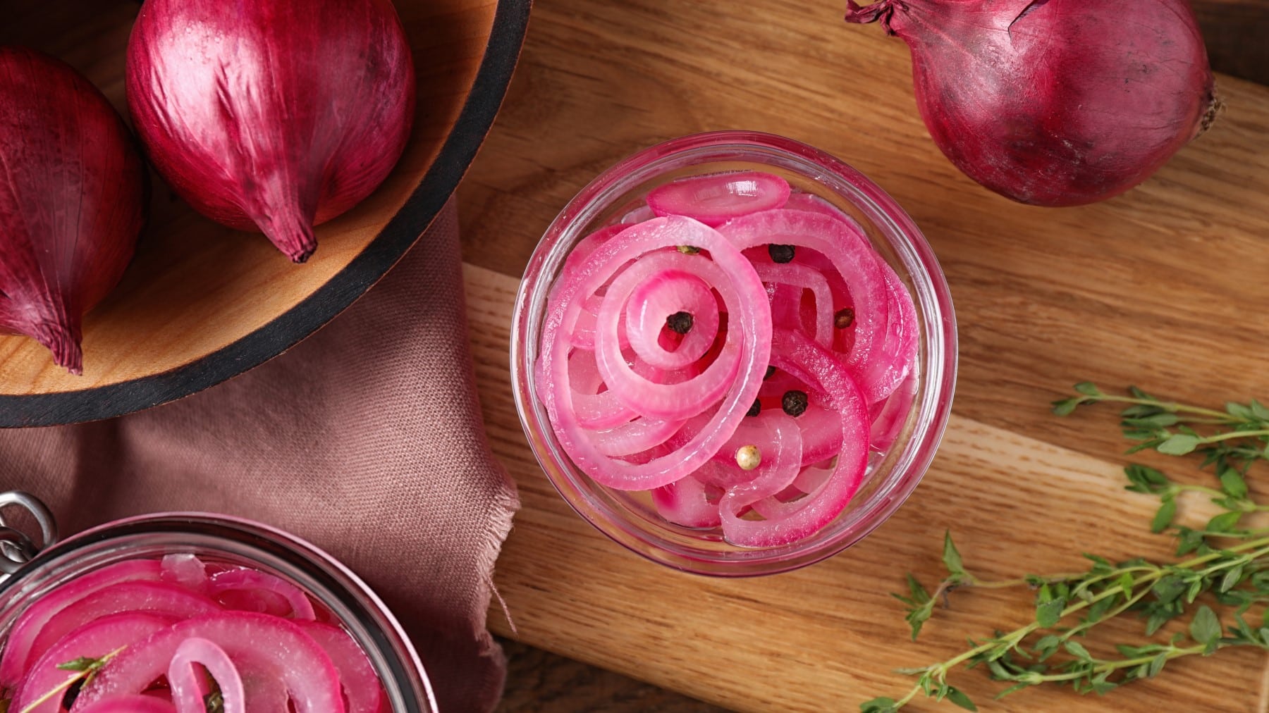 Pickled Red onions in einem Glas neben ganzen roten Zwiebeln und Thymianzweigen vor Holzhintergrund.