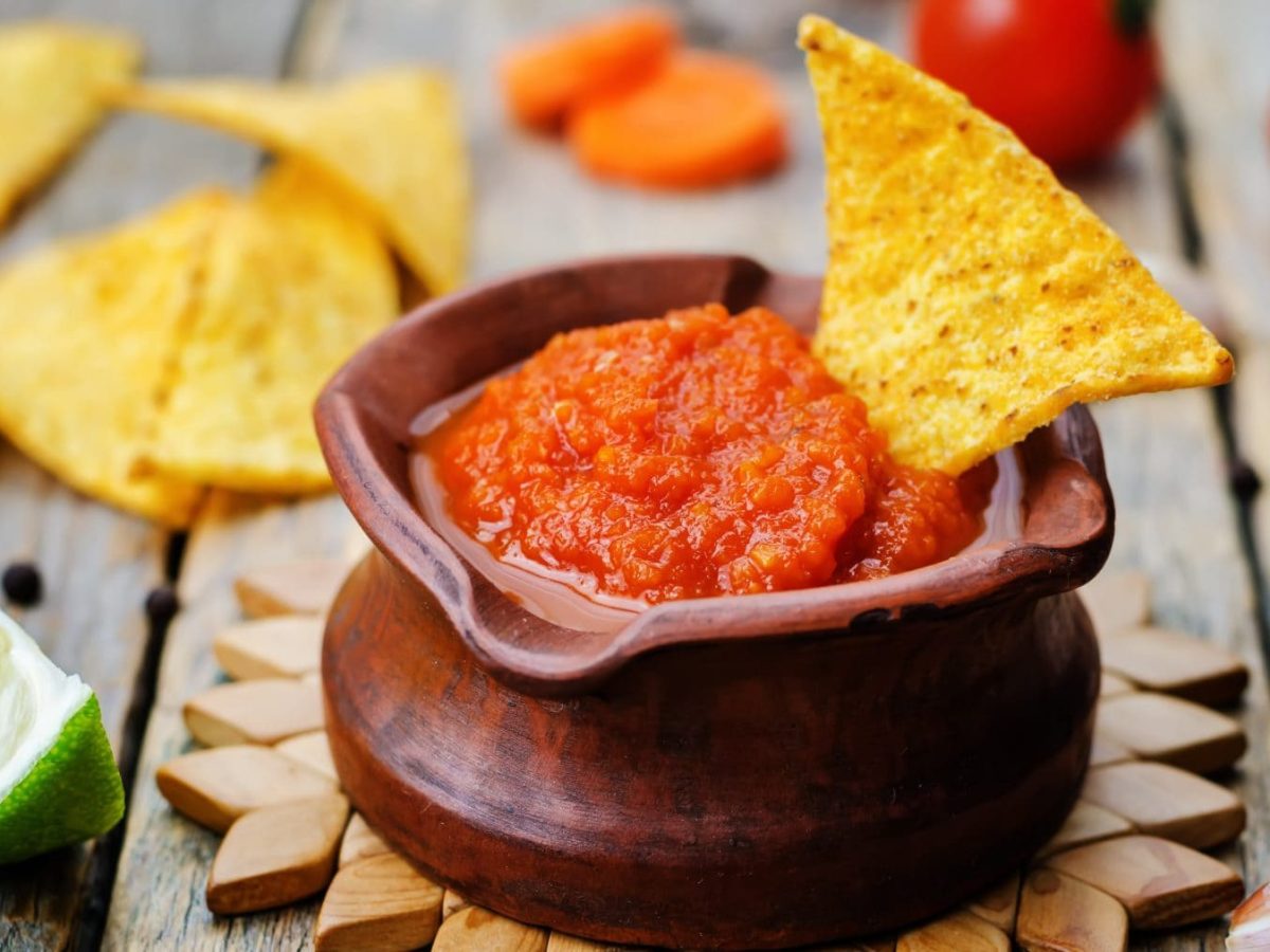 Salsa Taquera Roja in Tonschale auf Holzsternuntersetzer. Danaben Limette, Nachos und Tomaten. Frontalansicht.