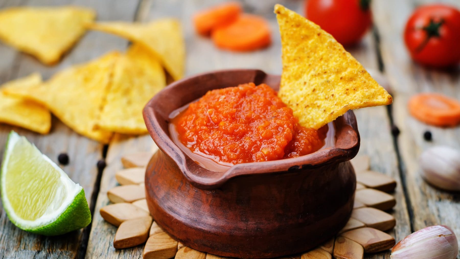 Salsa Taquera Roja in Tonschale auf Holzsternuntersetzer. Danaben Limette, Nachos und Tomaten. Frontalansicht.