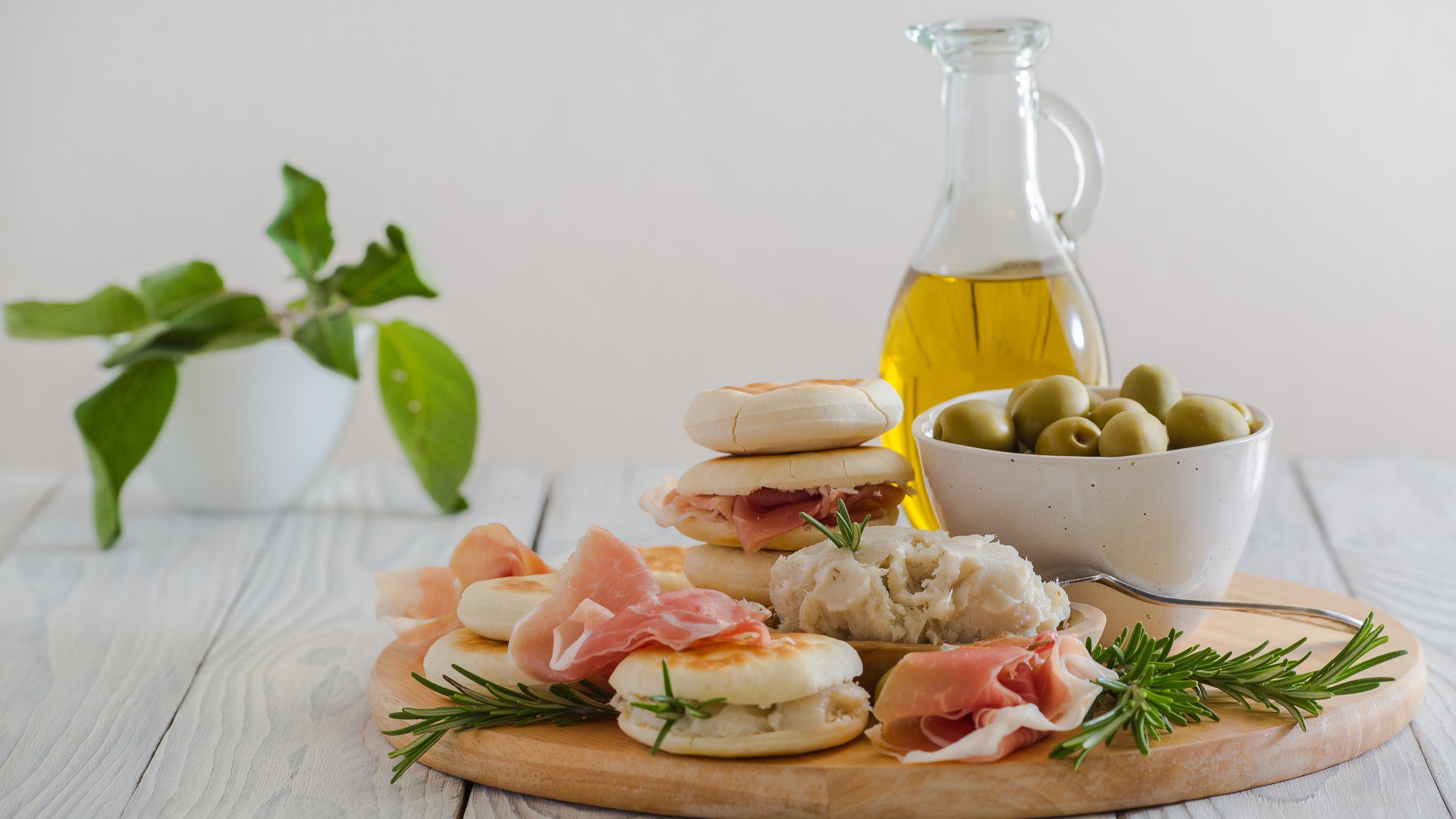Tigelle mit La Cunza auf Holzbrett mit rohem Schinken, Oliven in einer Schale und Rosmarin. Im Hintergrund Lorbeer und Olivenöl. Frontalansicht.