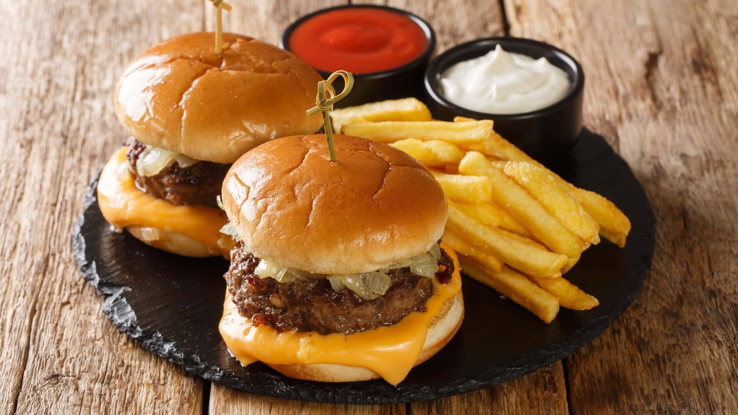 Zwei Wisconsin Butter Burger mit Pommes und je einer Schale Mayo und Ketchup auf Schieferplatte auf Holz. Draufsicht.