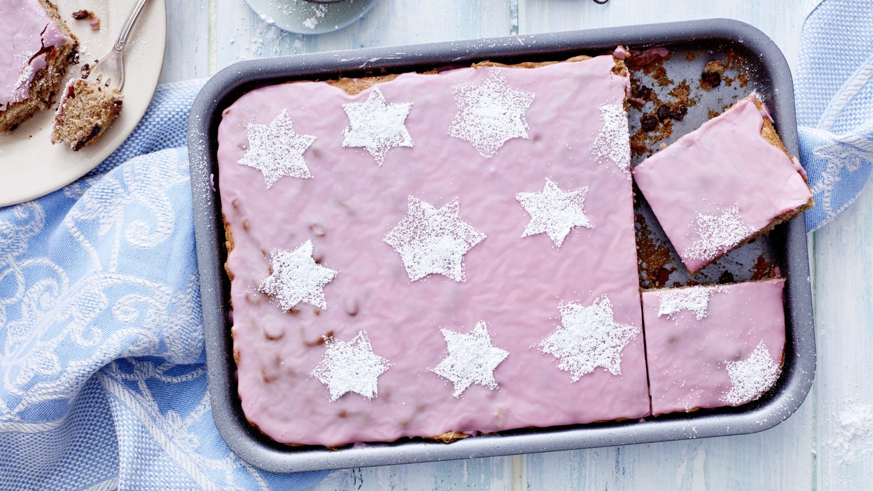 Glühweinkuchen vom Blech in der Form mit rosa Zuckerguss und Puderzuckersternen verziert vor blauem Hintergrund.