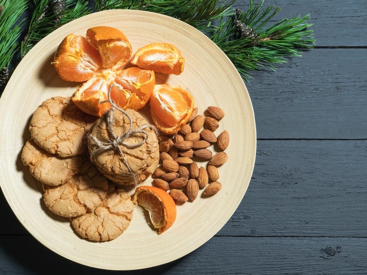 Draufsicht: Ein hölzerner Teller auf dem gestapelte Kekse, Mandeln und Orangenstücke liegen. Daneben liegt ein Seidenkieferzweig. Alles auf einem grau-blauen hölzernen Untergrund