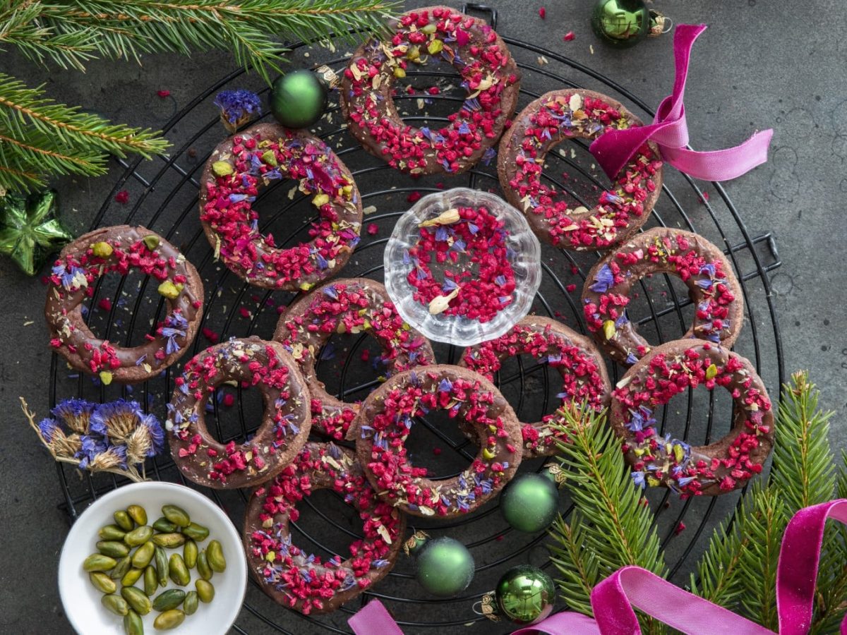 Mehrere Schokoladenkringel mit gefriergetrockneten Himbeeren auf einem Gitter neben Tannenzweigen, Pistazien, Weihnachtskugeln und pinkem Band vor grauem Hintergrund