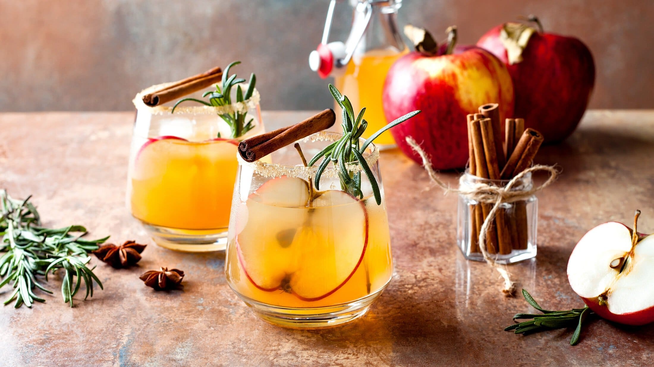 Zwei Gläser Apple Cider Margarita mit Rosmarin, Zimtstangen und Äpfeln.