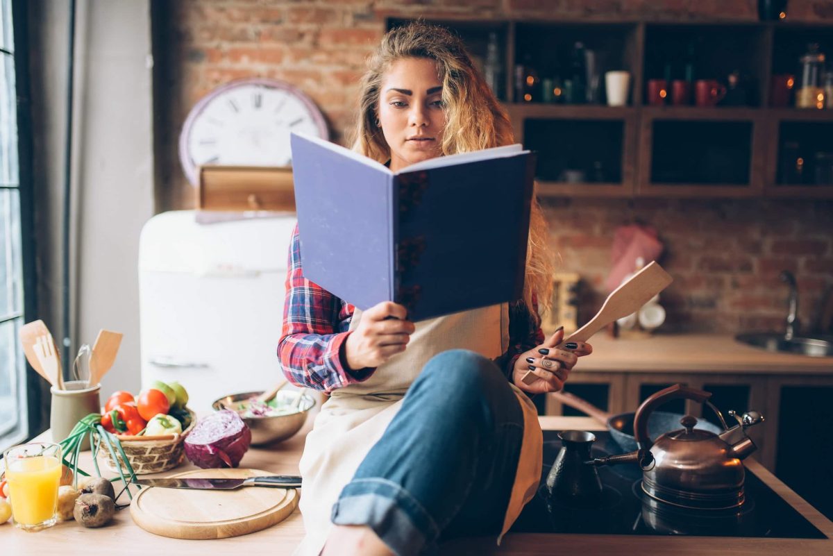 Deutscher Kochbuchpreis 2022: eine Frau sitzt mit einem Kochbuch in der Küche und liest darin.