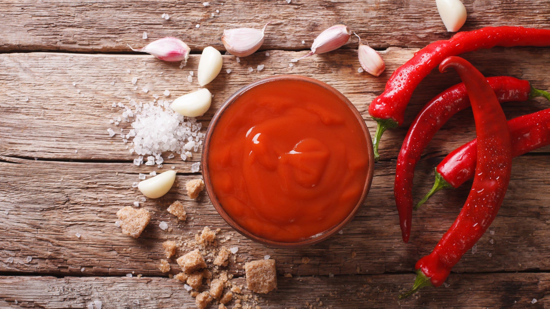 Selbst gemachte original Sriracha-Sauce aus Thailand in Schale auf Holz. Daneben rote Chilis, Knoblauch, grobes Salz und Palmzucker. Draufsicht.