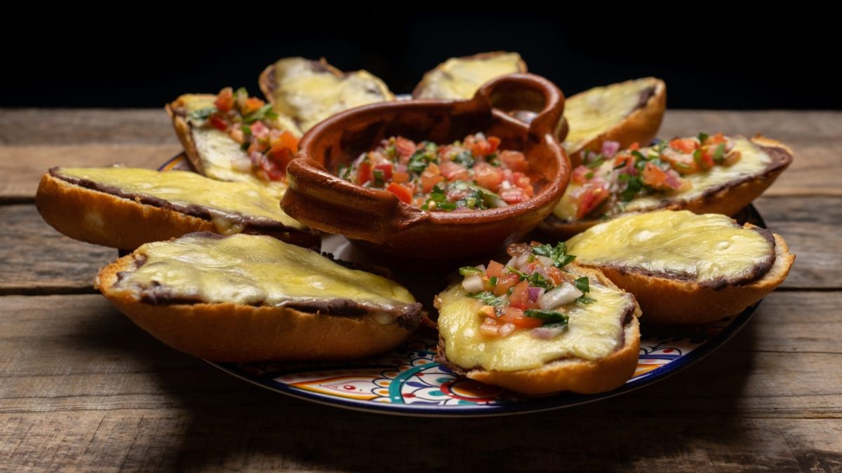 Molletes mit schwarzem Bohnenmus und Käse überbacken auf buntem Teller. In der Mitte eine Schale mit Pico de Gallo. Platziert auf Holz. Frontalansicht.