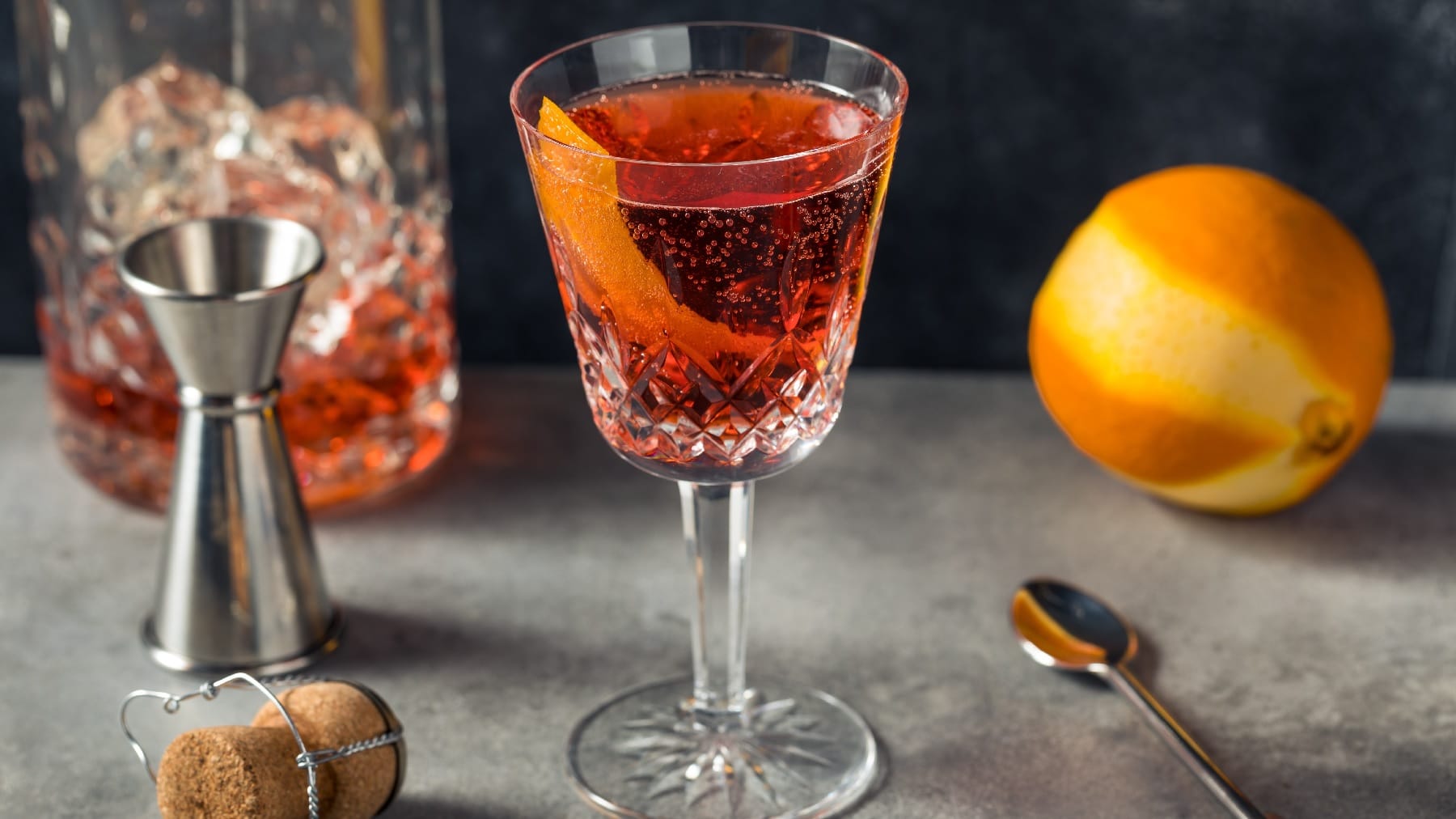 Ein Glas mit dem Negroni Sbagliato auf grauem Untergund mit einer angeschälten orange und einem Jigger.