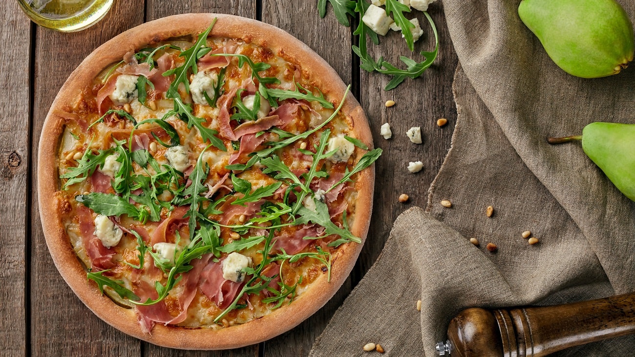 Draufsicht: Eine Pizza mit Schinken, Birne und Gorgonzola liegt auf einem Untergund aus Holz. Daneben liegen Birnen, Käsewürfel, Pinienkerne, eine Pfeffermühle und ein Tuch aus Leinen.