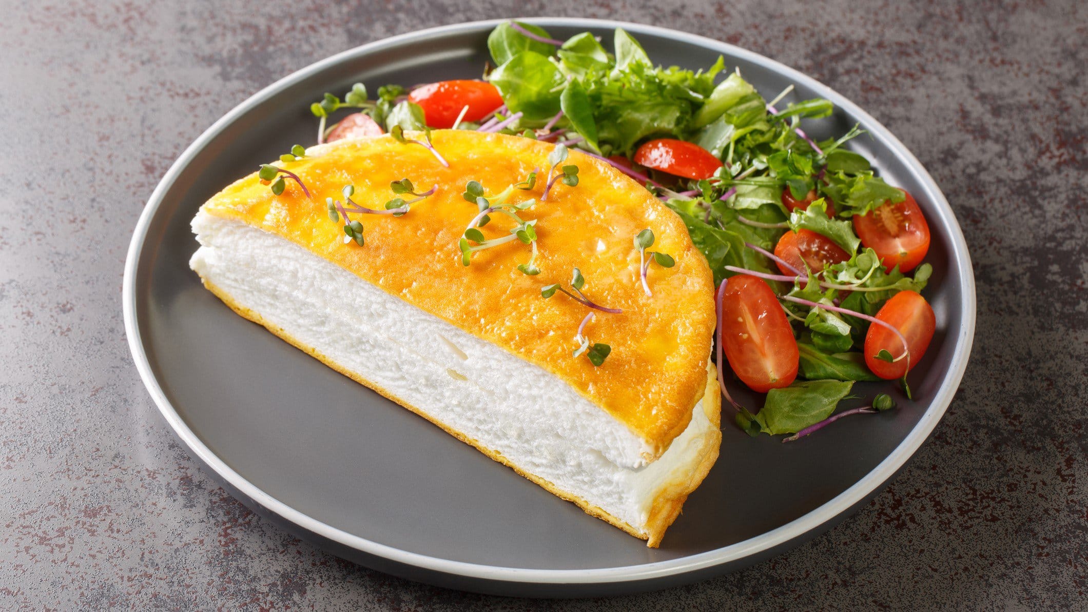 Ein Poulard-Omelett auf einem grauen Teller m und grauem Untergrund.it etwas Salat