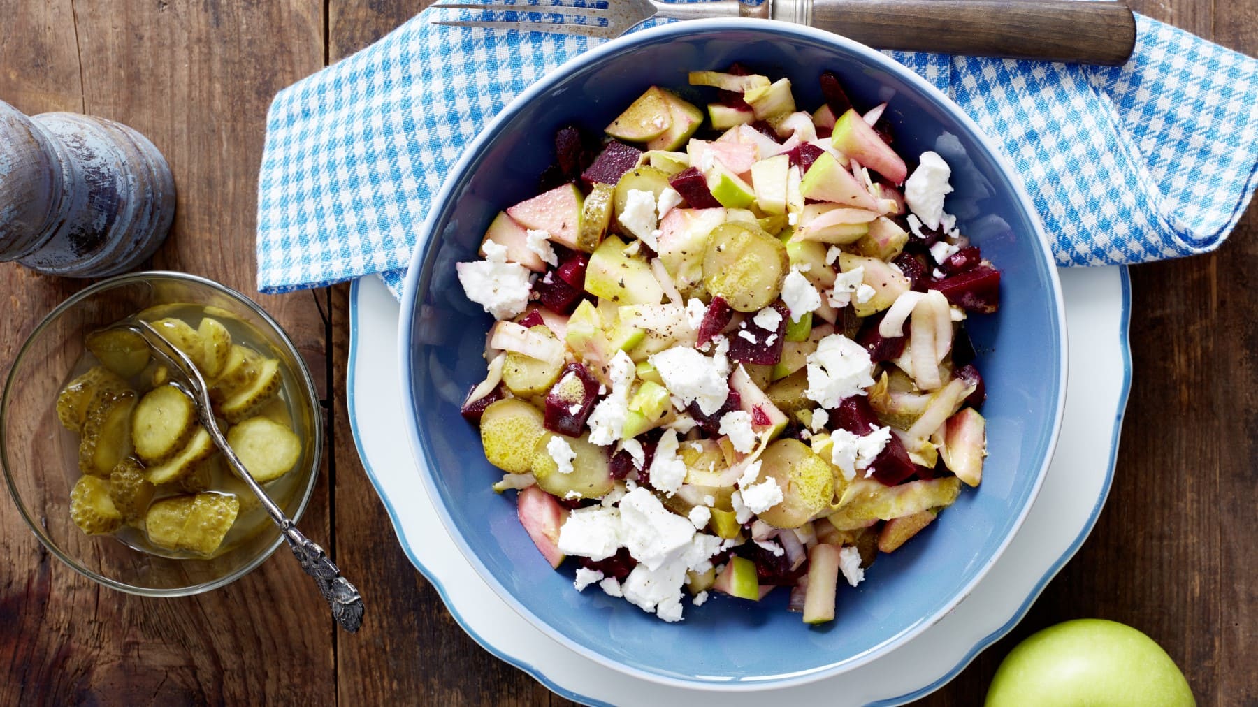 Rote-Bete-Salat mit Schafskäse in blauer Schüssel, daneben Gewürzmühle, saure Gurken und ein grüner Apfel vor Holzhintergrund.