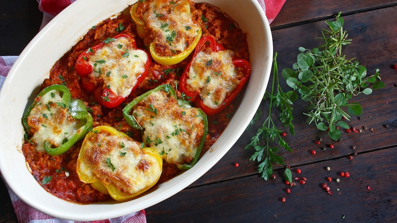 Draufsicht: Eine ofenfeste Form mit gefüllten Paprika-Hälften, die in einer Tomatensauce liegen. daneben liegen Kräuter.