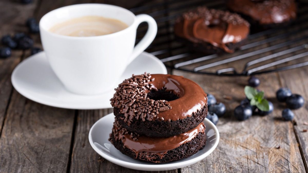 Triple Chocolate Donuts, bestehend aus Schokodunts mit Schokoglasur und Schokostreuseln, aufeinander gestapelt serviert mit einer Tasse Kaffee auf dunklem Holztisch.