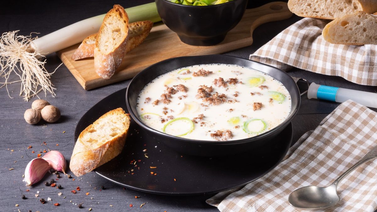Ein tiefer Teller vegane Käse-Lauch-Suppe mit Hack, daneben Baguette, Knoblauch, Muskatnüsse und Lauch.