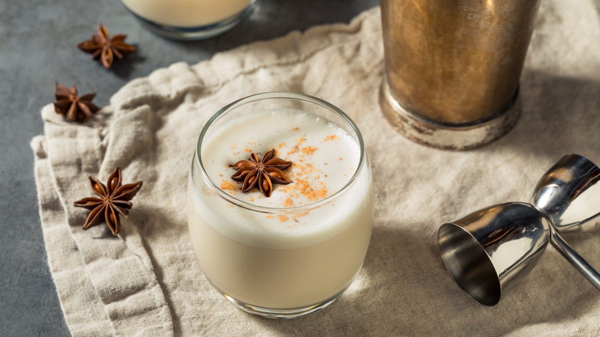 Vanilla Maple Bourbon Milk Punch im Glas mit Sternanis, Barmaß und Shaker auf Tuch und grauem Untergrund. Im Hintergrund ein weiteres Glas. Draufsicht.