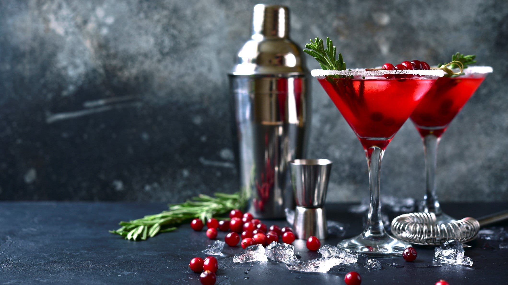 Zwei Christmas-Margarita in Cocktailschale mit Tannenzweig. Im Bild Shaker, Barmaß, Cranberrys und Zweige verteilt. Frontalansicht.