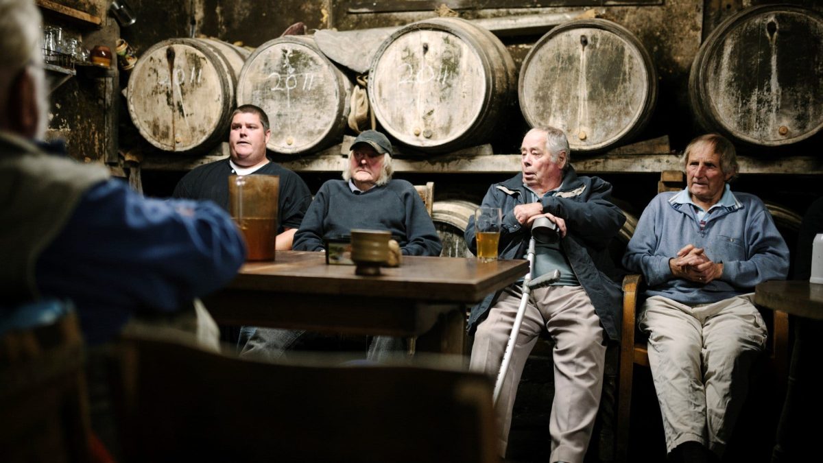 Was sind die Unterschiede zwischen Cidre, Cider und Apfelwein? Vier alte Männer vor Holzfässern an einem Tisch mit Tassen und Flaschen. Frontalansicht.