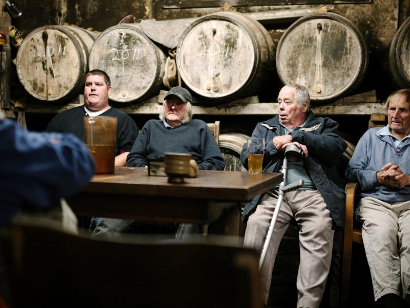 Was sind die Unterschiede zwischen Cidre, Cider und Apfelwein? Vier alte Männer vor Holzfässern an einem Tisch mit Tassen und Flaschen. Frontalansicht.