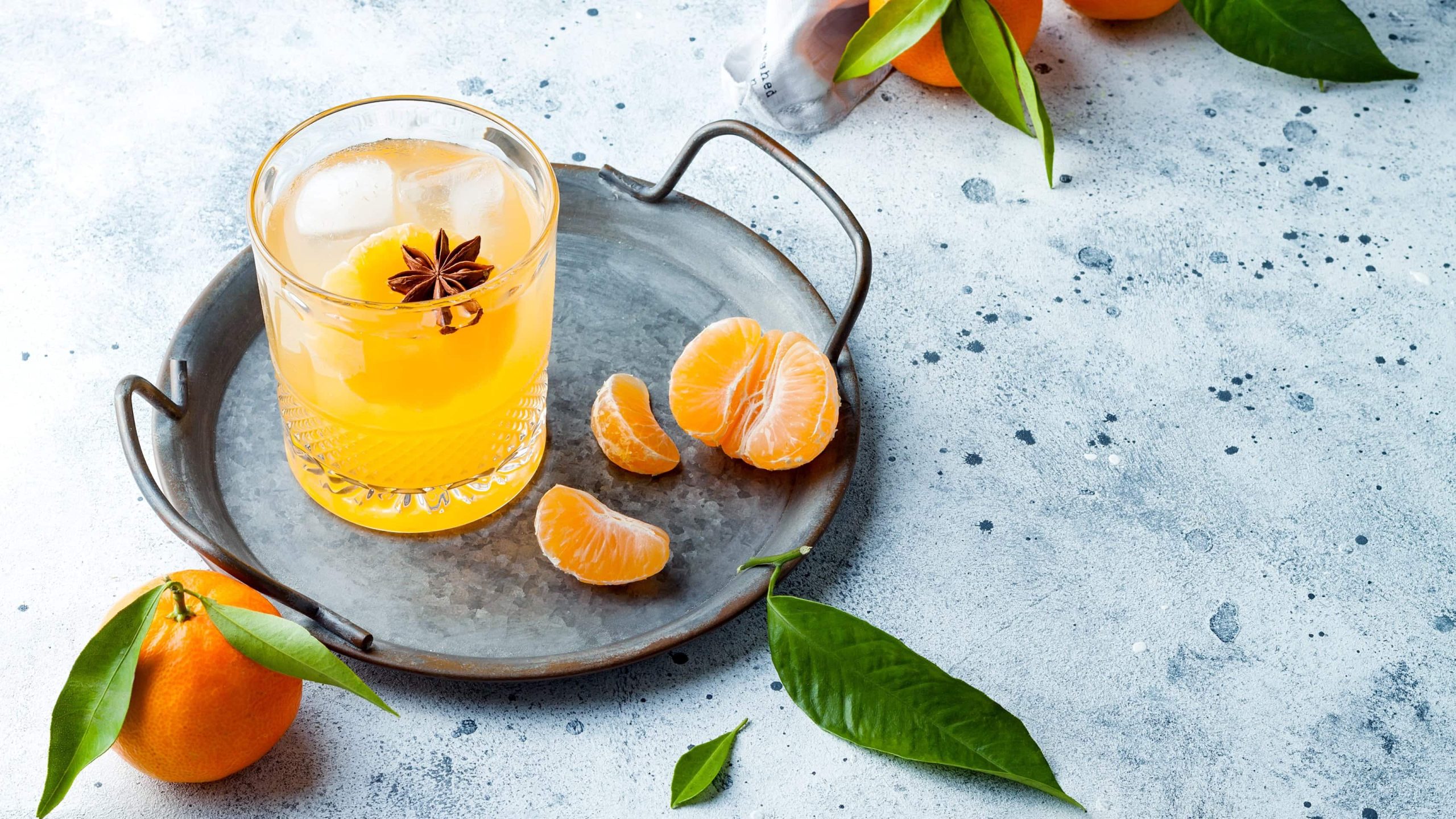 Ein graues Tablett mit Mandarinen-Cocktail mit Ingwer neben Mandarinenstücken.