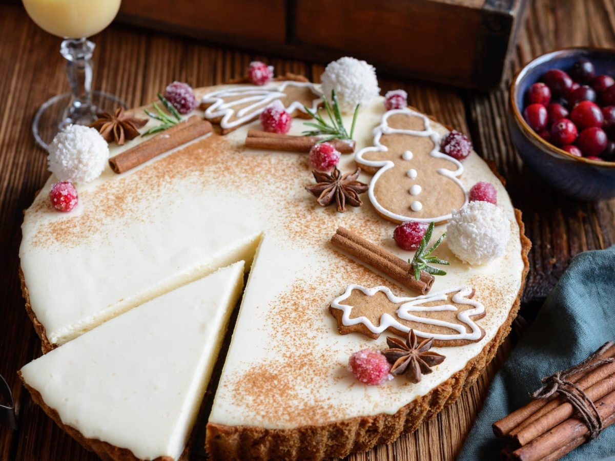 Eggnog Cheesecake mit kandierten Preiselbeeren und Spekulatiusboden auf Holztisch. Dekoriert mit Weihnachtsgebäck. Im Bild ein Glas Eierlikör, Gewürze und Preiselbeeren. Draufsicht.