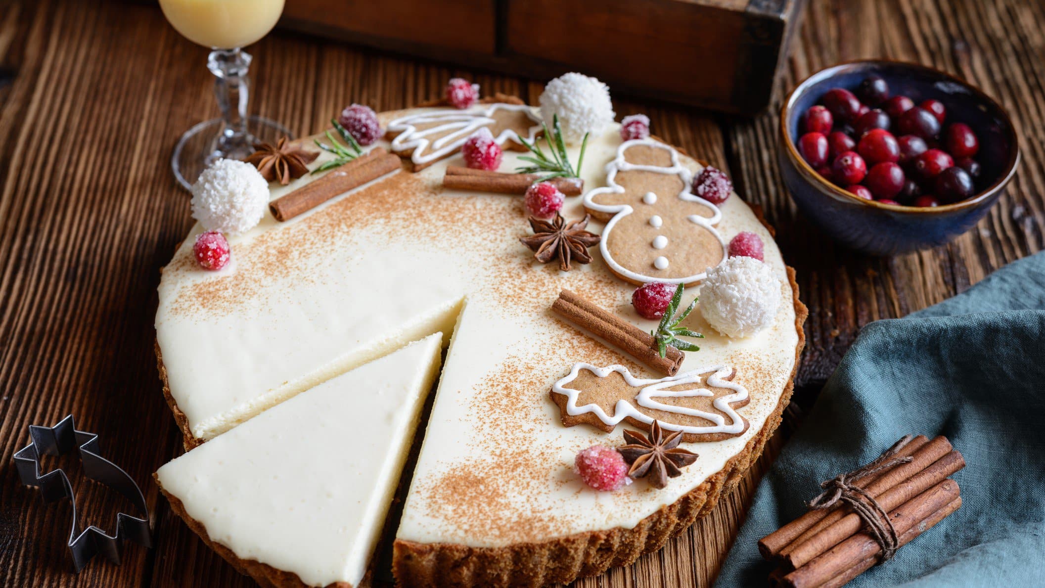 Eggnog Cheesecake mit kandierten Preiselbeeren und Spekulatiusboden auf Holztisch. Dekoriert mit Weihnachtsgebäck. Im Bild ein Glas Eierlikör, Gewürze und Preiselbeeren. Draufsicht.