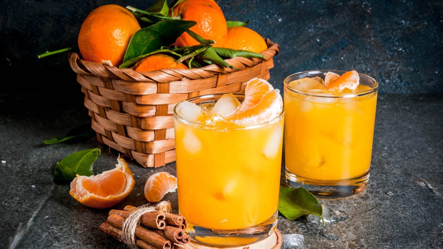 Mandarinen Cocktail mit Vodka, Zimt und Anis in zwei Gläsern mit Eis. Dahinter eine Schale mit Mandarinen. Im Bild Mandarinenstücke und Zimtstangen. Frontalansicht.