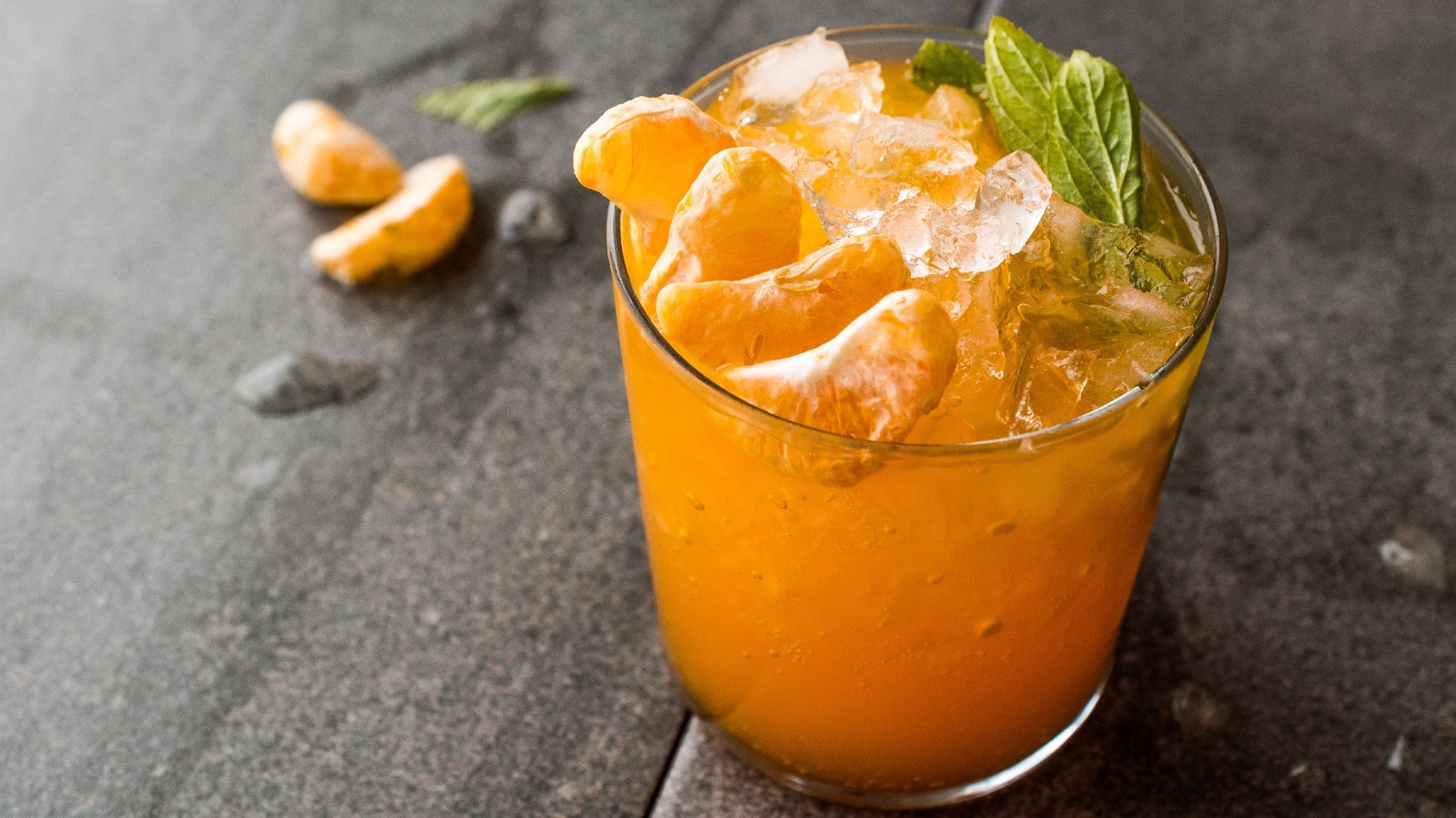 Ein Glas mit dem Mandarinen-Mojito auf einem grauen Tisch mit zwei Mandarinenstückchen und Eiswürfeln daneben.