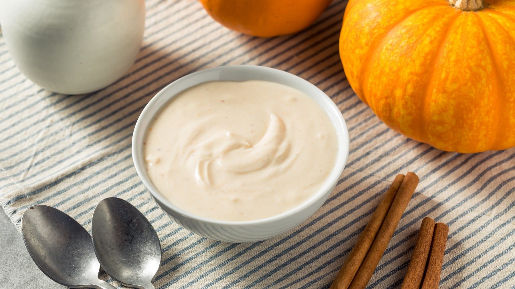 Pumpkin Spice Joghurt, serviert in einer Schale auf einem einfach gedeckten Küchentisch mitsamt ein paar Stangen Zimt und einem Hokkaidokürbis.