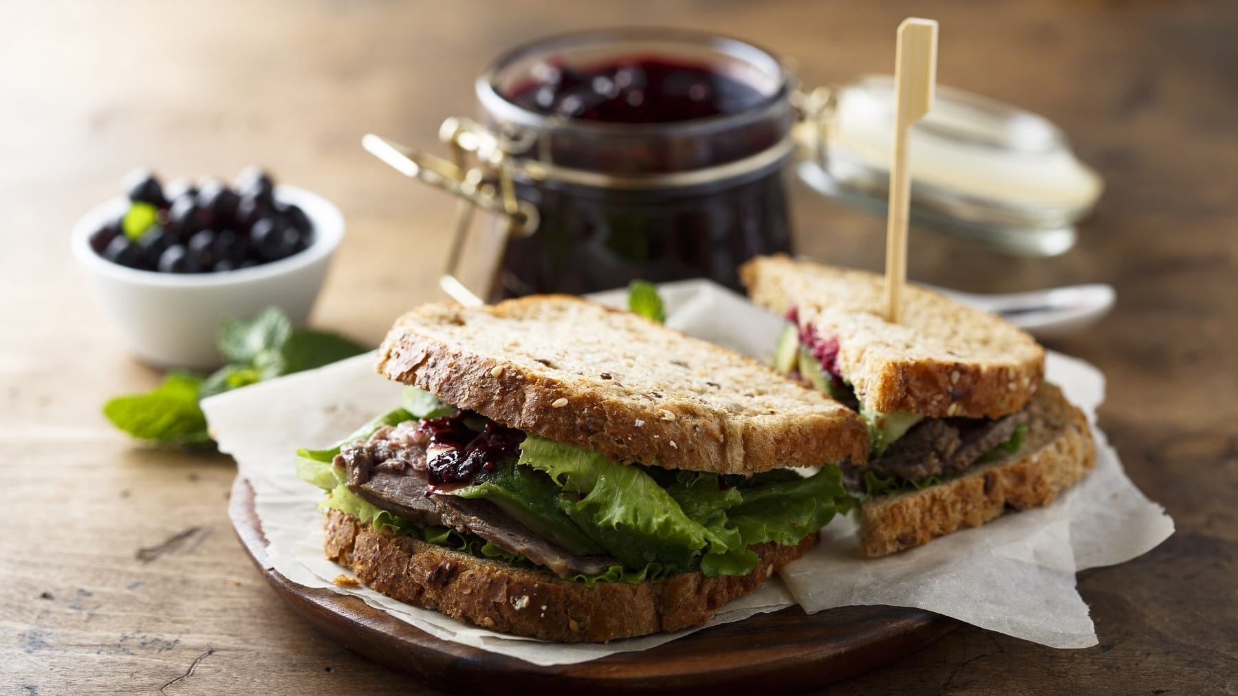 Roastbeef-Sandwich mit Heidebeermarmelade und knackigem Salat, aufgeschnitten serviert auf einem Teller mit noch mehr Marmelade auf einem Holztisch.