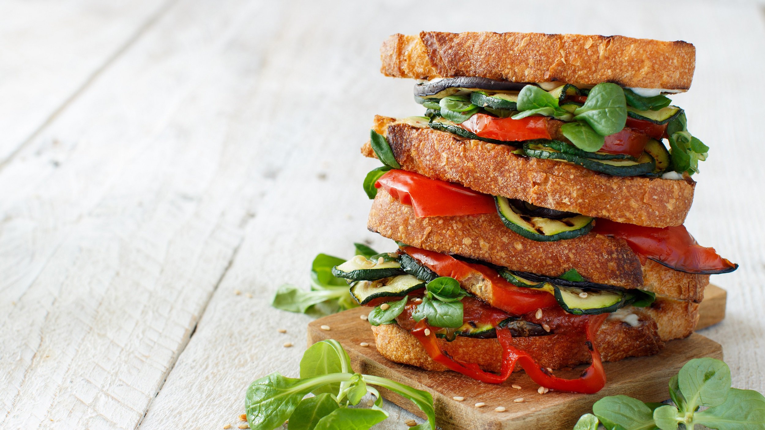 Ein gestapeltes Sandwich mit Grillgemüse, Feldsalat und Tomaten auf hellem Holz.