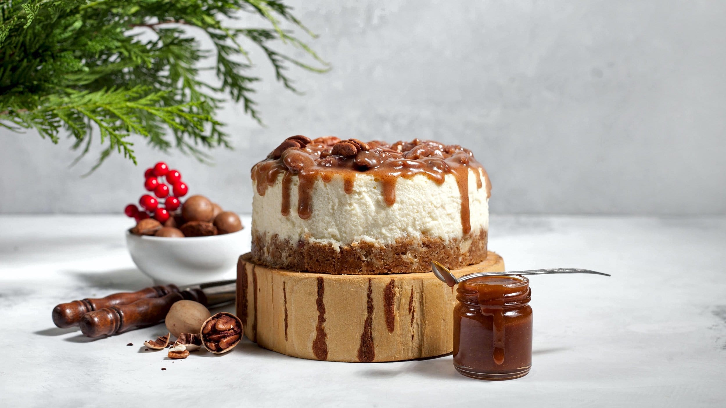 Ein Spekulatius-Cheesecake auf einem Stück Holz, daneben ein Glas mit Karamellsauce und Weihnachtsdeko.