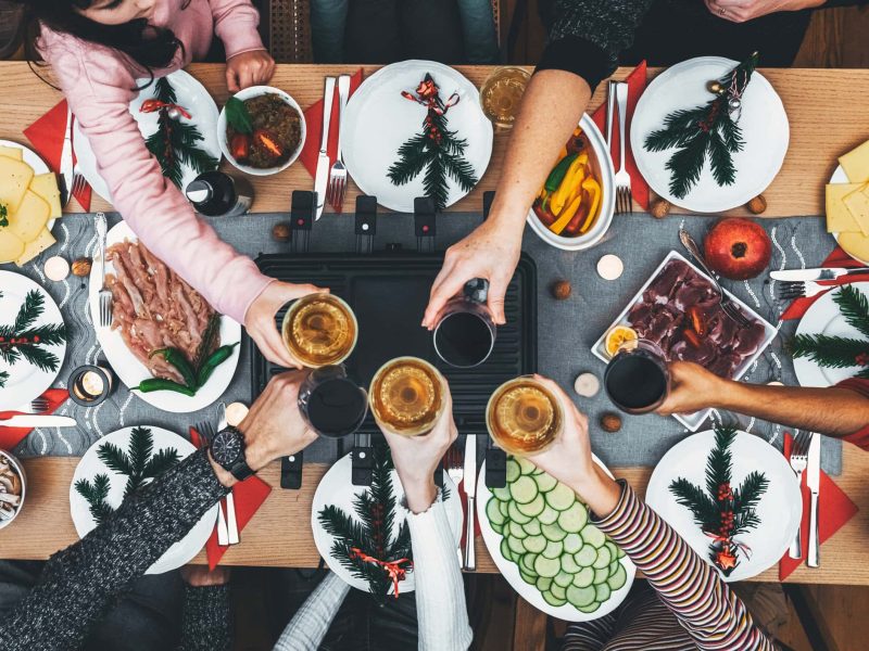 Veganes Raclette: Eine Dinnergesellschaft an einem Tisch, auf dem ein Raclette aufgebaut ist, alle stoßen mit Getränken an. Draufsicht.